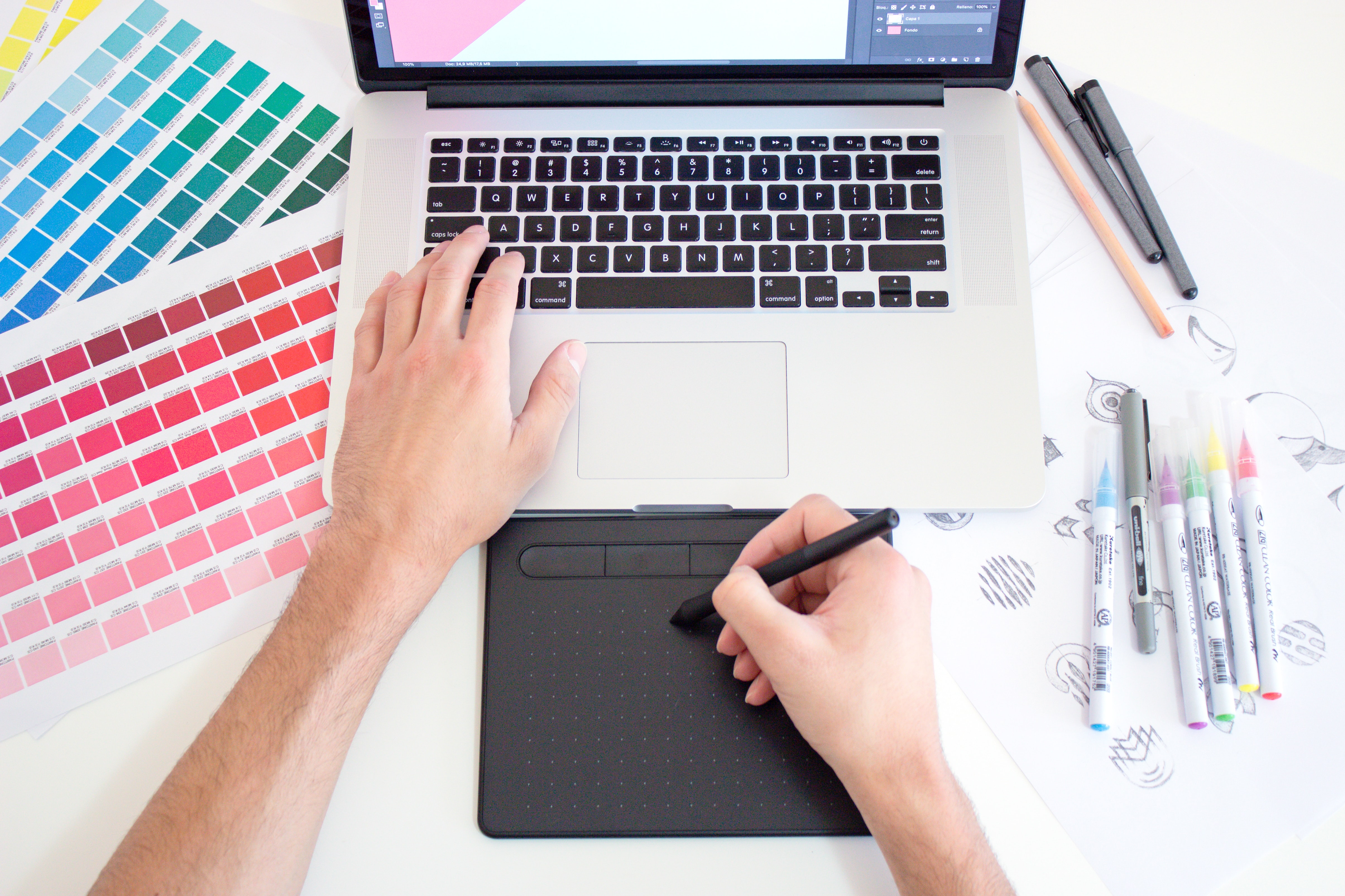 A person working with a graphics tablet on a laptop. Around the laptop are color charts, felt-tip pens and sketches.